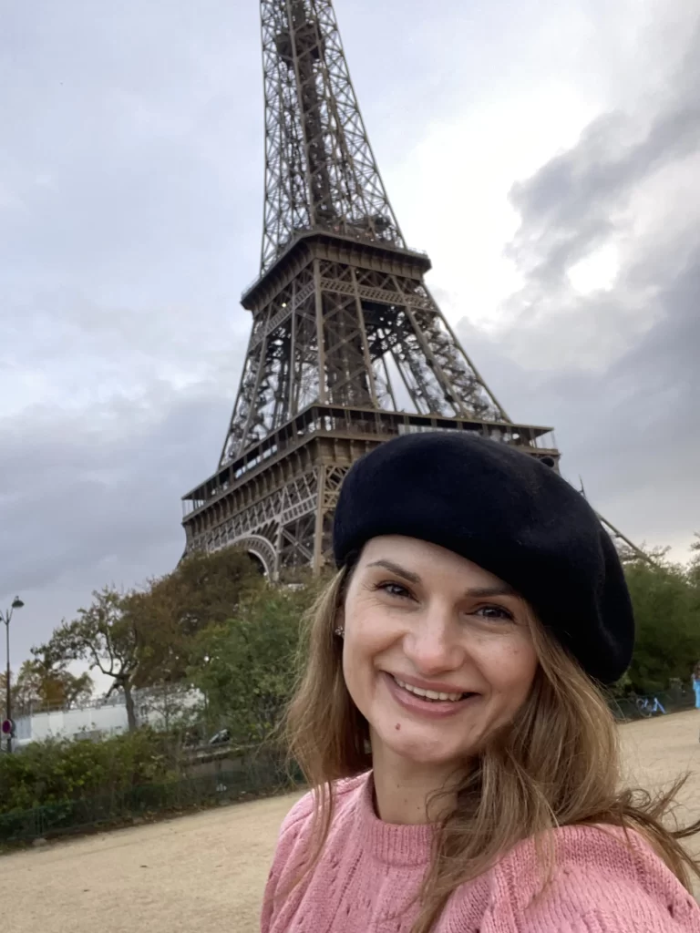 Femeie zâmbitoare purtând un beret negru, pozând în fața Turnului Eiffel într-o zi înnourată la Paris.