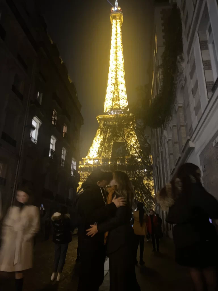 Cuplu sărutându-se romantic în fața Turnului Eiffel luminat pe timp de noapte, într-o stradă pariziană aglomerată