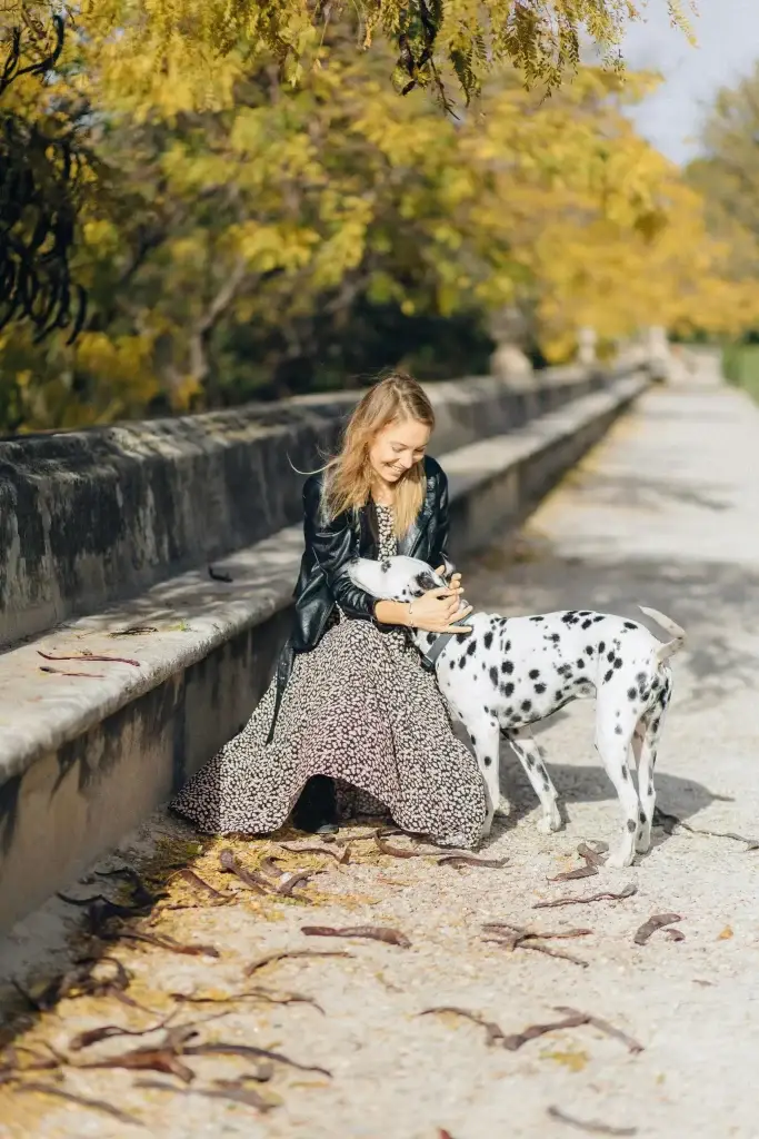 O femeie îmbracată într-o rochie lungă cu animal print și o geacă din piele neagră, mângâind un câine dalmațian